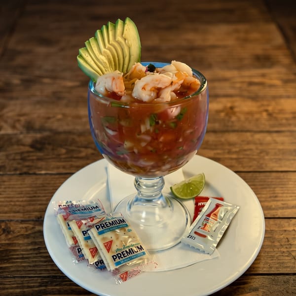 Shrimp cocktail in a glass with avocado, lime, and cracker packets on a white plate.