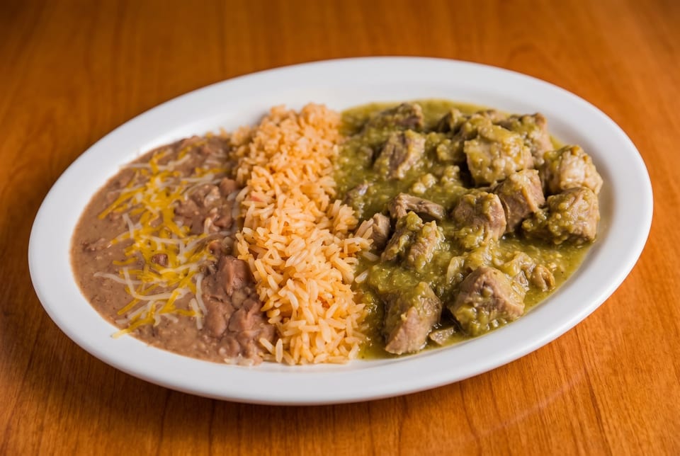 A white oval plate with chile verde, Spanish rice, and refried beans topped with cheese.