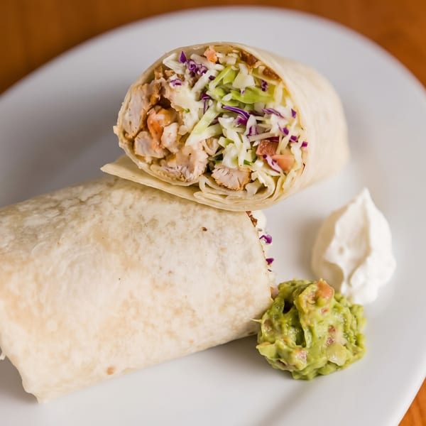Two chicken burritos, one cut open, with guacamole and sour cream on a white plate.