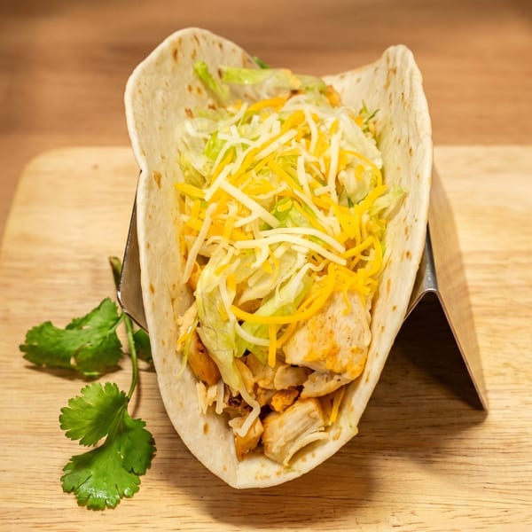 A chicken taco with shredded lettuce and cheese in a soft tortilla, on a wooden board.