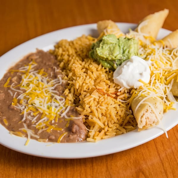 A white plate with refried beans, yellow rice, guacamole, sour cream, and taquitos.