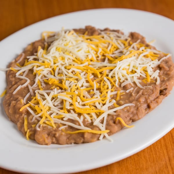 Refried beans topped with shredded yellow and white cheese on a white oval plate.