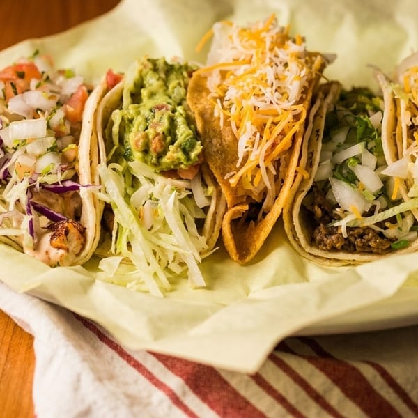 Five assorted tacos with various fillings like guacamole, cheese, and pico de gallo on a yellow paper.